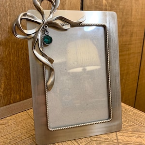 Könnte beinhalten: Silberfarbener rechteckiger Bilderrahmen mit dekorativer Schleife. Die Schleife hat fließende Bänder und einen hängenden teal-farbenen Schmuck. Der Rahmen hat einen seilartigen Rand um den Fotobereich.