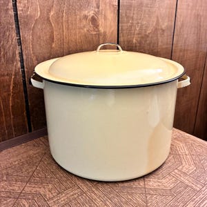 May include: A large, cylindrical, cream-coloured enamel stockpot with a matching lid. The pot features two side handles and a thin black rim around the top edge of the vessel and the lid. The surface has a smooth, glossy finish throughout.