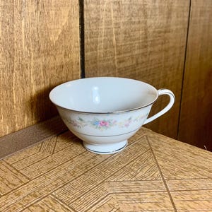 Op de afbeelding: Een wit theekopje met een bloemenpatroon in roze, blauw en geel. Het kopje heeft een zilveren rand en een zilveren band rond het midden. Het kopje staat op een houten oppervlak.