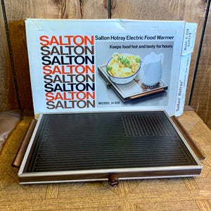Puede incluir: Un calentador de alimentos eléctrico con la marca "Salton" en la caja. El calentador tiene una superficie de calentamiento negra con asas de madera y un mando. La caja dice "Salton Hotray Electric Food Warmer" y "Keeps food hot and tasty for hours."