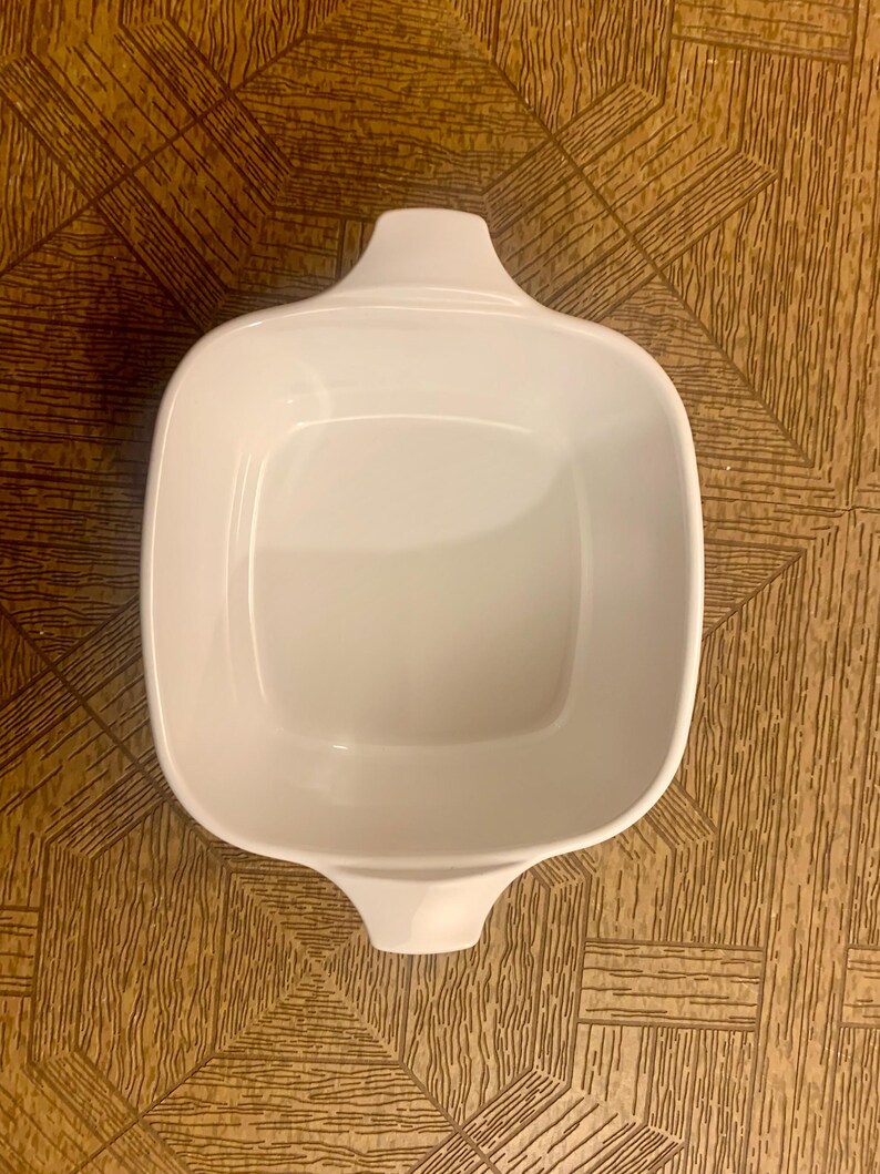 May include: White ceramic baking dish with two handles. The dish is square shaped and has a smooth, glossy finish.