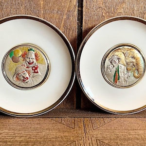 May include: Two white decorative plates with gold rims. Each plate features a central medallion with a circus theme. One plate shows two clowns, and the other shows a clown with an elephant. The plates are set against a wooden background.
