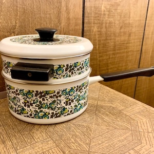 May include: White enamel double boiler with a black handle and lid knob. The pot features a floral pattern in shades of green, blue, and brown. The handle is approximately 6 inches long. The pot is sitting on a wooden surface.