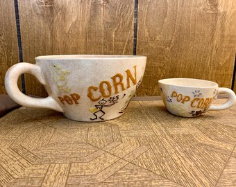 Op de afbeelding: Twee keramische popcornmokken, een grote en een kleine, met handvatten. De mokken zijn beige met bruine letters met de tekst "POP CORN". De grote mok heeft een stokfiguurontwerp. De kleine mok heeft bloemmotieven.