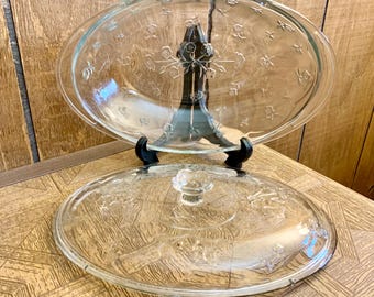May include: Clear glass oval casserole dish with lid. The lid has a clear knob and floral design. The dish has a floral design and is supported by a black metal stand. The dish is on a wooden surface.