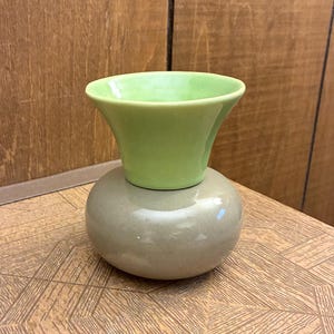May include: A two-toned ceramic vase featuring a wide, light green flared top section and a rounded, smooth taupe-colored base. The vessel has a glossy finish and a distinct separation between the upper funnel-shaped opening and the bulbous bottom.