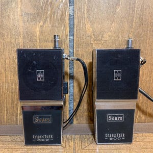 May include: Two vintage Sears transTalk 1000 walkie-talkies. Each unit has a black speaker grill, a silver antenna, and a lower section with the Sears logo and the transTalk model name. The units are connected by a black cable.