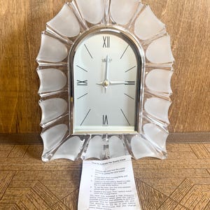 May include: A clear quartz clock with a silver frame and a decorative, frosted glass border. The clock face is white with black Roman numerals and hands. A small paper with instructions is attached to the clock.