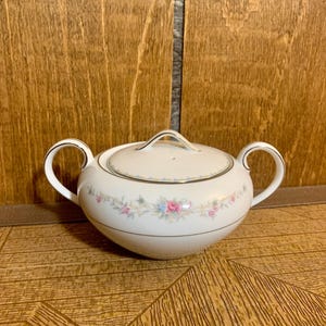 Puede incluir: Azucarero de porcelana blanca con tapa y dos asas. El tazón tiene un diseño floral con flores rosas, azules y verdes. La tapa tiene un borde dorado.