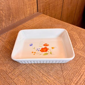 May include: A rectangular white ceramic baking dish with a fluted exterior edge. The interior base features a colorful floral illustration with orange, red, blue, and yellow flowers and green stems centered on the smooth white surface.