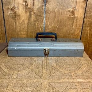 Peut inclure: Une boîte à outils en métal gris avec une poignée en plastique noir. La boîte à outils a un loquet sur le devant et est légèrement patinée.