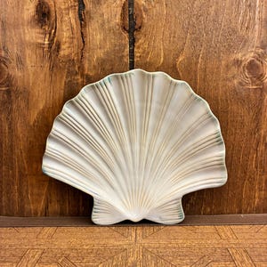 Könnte beinhalten: Eine Keramikschale in Form einer fächerartigen Muschel mit glänzendem cremeweißem Finish und dezenten bläulich-grünen Akzenten entlang der gewellten Ränder und strahlenförmigen Rippen, die von der Basis bis zum äußeren Rand verlaufen.
