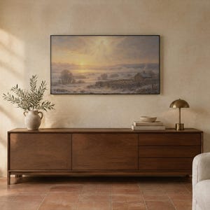 May include: A rectangular landscape painting featuring a misty sunrise over a rural field with a small house, stone walls, and a faint winged figure in the sky, displayed in a thin black frame above a long wooden sideboard with drawers.