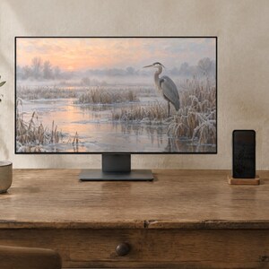 May include: A computer monitor displaying a landscape painting of a heron standing in a frosty marsh at sunrise. The monitor has a thin black frame and a dark gray stand, positioned on a wooden desk next to a smartphone and a ceramic mug.