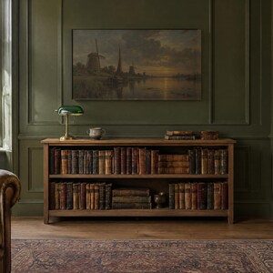 May include: A wooden bookcase with two shelves filled with antique books. On top sits a green glass banker lamp and a small ceramic bowl. Above the bookcase hangs a landscape painting of windmills by a river on a dark olive green wall.
