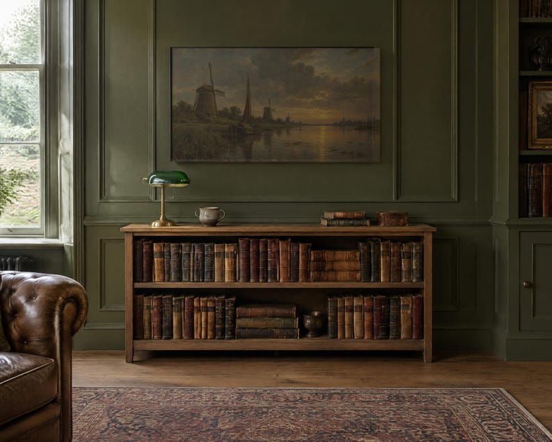 May include: A wooden bookcase with two shelves filled with antique books. On top sits a green glass banker lamp and a small ceramic bowl. Above the bookcase hangs a landscape painting of windmills by a river on a dark olive green wall.