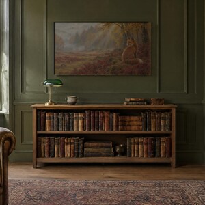 May include: A wooden bookshelf with two tiers filled with antique books, topped with a green glass banker lamp and a small ceramic bowl. Above the shelf hangs a landscape painting of a fox in a forest, set against a dark olive green wall.