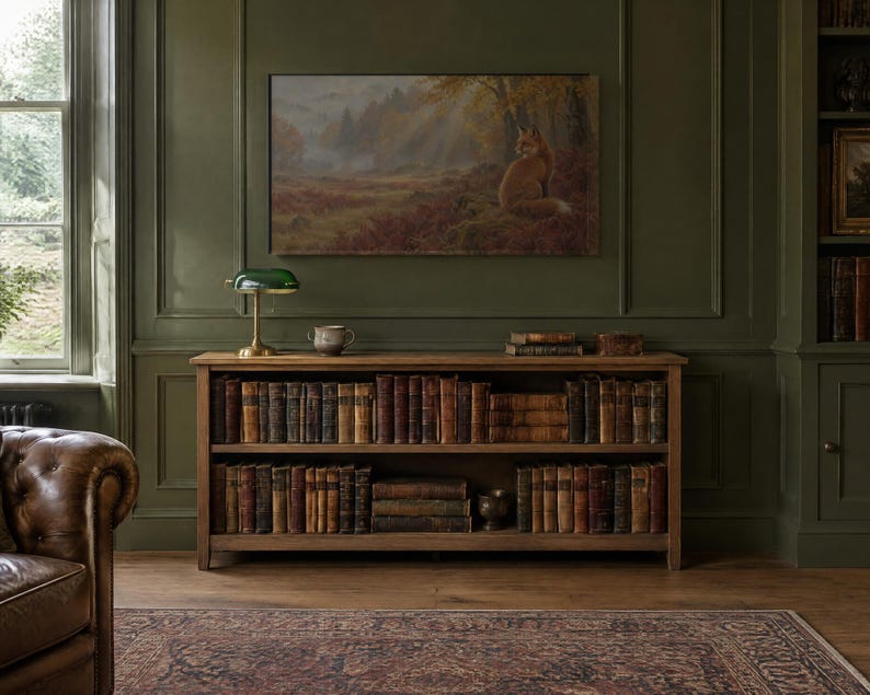 May include: A wooden bookshelf with two tiers filled with antique books, topped with a green glass banker lamp and a small ceramic bowl. Above the shelf hangs a landscape painting of a fox in a forest, set against a dark olive green wall.