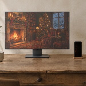 May include: A computer monitor displaying a cozy room with a fireplace and a Christmas tree, placed on a wooden desk next to a ceramic mug, a smartphone on a wooden stand, and a notebook. The monitor has a thin black frame and a dark base.