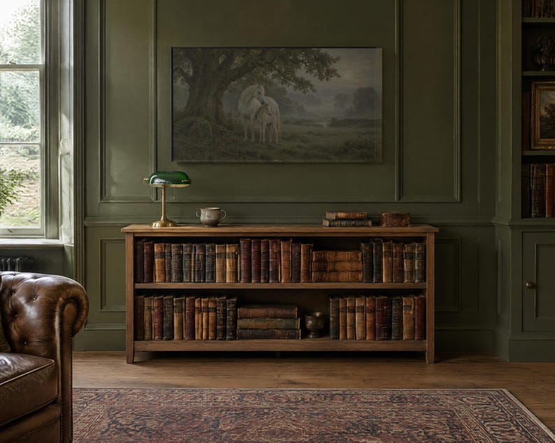 May include: A wooden bookshelf with two shelves filled with antique books, topped with a green glass banker lamp and a small ceramic bowl. The unit is positioned against a dark olive green paneled wall beneath a framed landscape painting.