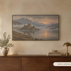 May include: A rectangular landscape painting in a thin black frame depicting a stone castle on a small island in a lake surrounded by misty mountains at sunset. The artwork features soft golden light reflecting on the water and cloudy skies.