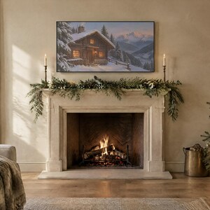 May include: A rectangular television screen mounted above a stone fireplace displays a winter landscape painting of a wooden cabin in the snow. The fireplace mantel is decorated with green pine garland and two tall, thin metal candle holders.