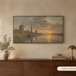 May include: A framed landscape painting depicting a river scene with windmills and a sailboat under a sunset sky. The artwork is displayed on a wall above a wooden sideboard, which holds a ceramic vase with greenery, books, and a small lamp.