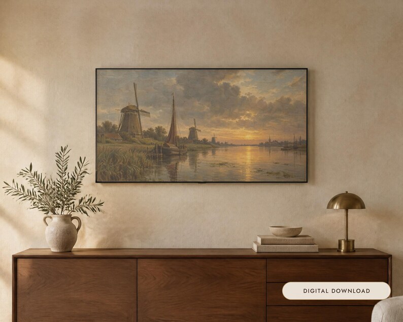 May include: A framed landscape painting depicting a river scene with windmills and a sailboat under a sunset sky. The artwork is displayed on a wall above a wooden sideboard, which holds a ceramic vase with greenery, books, and a small lamp.