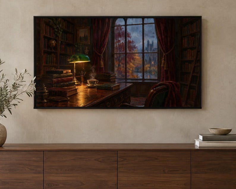 May include: A framed wall art print depicting a dark, wood-paneled library interior with a green banker lamp illuminating a desk, stacks of books, a steaming teacup, and a large window overlooking an autumn landscape with red trees.