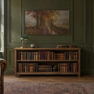 May include: A wooden bookcase with two shelves filled with antique books, topped with a green glass banker lamp and a small ceramic bowl. A landscape painting of an autumn forest path hangs on the dark green paneled wall above the unit.