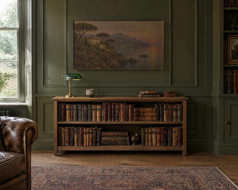 Op de afbeelding: Een houten boekenkast met twee planken gevuld met rijen antieke boeken. Op de bovenste plank staat een groene glazen bankierslamp en een kleine keramische kom. Een landschapsschilderij hangt aan de olijfgroene muur boven de kast.