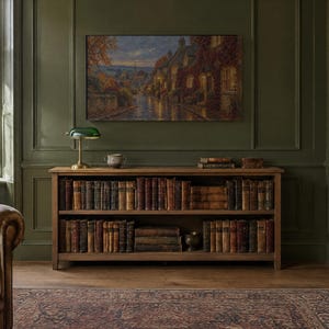 K&ouml;nnte beinhalten: Ein h&ouml;lzernes B&uuml;cherregal mit zwei Ebenen voller antiker B&uuml;cher, gekr&ouml;nt von einer gr&uuml;nen Bankerlampe aus Glas und einer kleinen Keramikschale. &Uuml;ber dem Regal h&auml;ngt ein Landschaftsgem&auml;lde einer Dorfstra&szlig;e in der Abendd&auml;mmerung mit warm leuchtenden Fenstern.