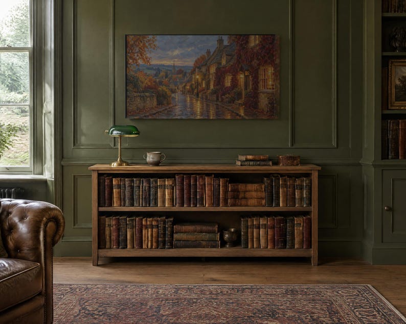 K&ouml;nnte beinhalten: Ein h&ouml;lzernes B&uuml;cherregal mit zwei Ebenen voller antiker B&uuml;cher, gekr&ouml;nt von einer gr&uuml;nen Bankerlampe aus Glas und einer kleinen Keramikschale. &Uuml;ber dem Regal h&auml;ngt ein Landschaftsgem&auml;lde einer Dorfstra&szlig;e in der Abendd&auml;mmerung mit warm leuchtenden Fenstern.