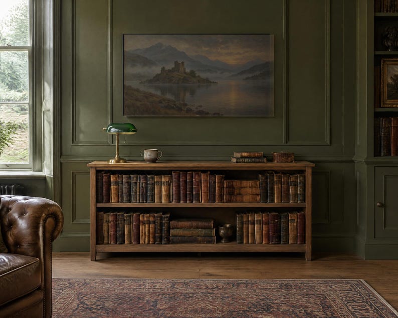 May include: A wooden bookshelf with two shelves filled with antique books, positioned against a dark green paneled wall. A green banker lamp and a small ceramic bowl sit on top of the shelf, beneath a framed landscape painting of a castle.
