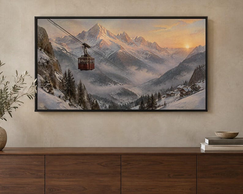 May include: A framed landscape painting depicting a red cable car suspended over a snow-covered mountain valley at sunset. The scene features jagged, snow-capped peaks, evergreen trees, and small cabins nestled in the misty, wintry terrain.