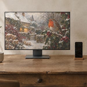 May include: A computer monitor displaying a winter scene with a robin perched on a holly bush in front of a snow-covered cottage. The monitor has a thin black frame and a dark gray stand, positioned on a wooden desk with a mug and a smartphone.