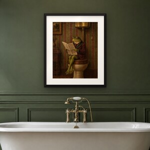 May include: A framed wall art print featuring a green frog wearing a dark red jacket, sitting on a toilet and reading a newspaper. The scene is set in a room with striped wallpaper, rendered in muted tones of brown, green, and cream.