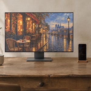 May include: A computer monitor displaying a painting of a Parisian street scene with a cafe, wet cobblestone streets, and the Notre Dame cathedral in the distance. The monitor has a thin black frame and a dark gray stand, positioned on a wooden desk.