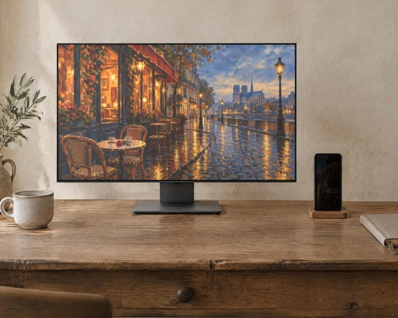 May include: A computer monitor displaying a painting of a Parisian street scene with a cafe, wet cobblestone streets, and the Notre Dame cathedral in the distance. The monitor has a thin black frame and a dark gray stand, positioned on a wooden desk.