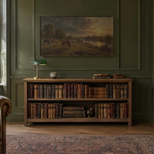 May include: A wooden two-shelf bookcase filled with antique books, topped with a green glass banker's lamp and a small ceramic bowl. Above the bookcase hangs a landscape painting of horses grazing in a field at sunset.
