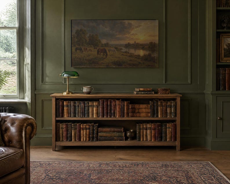 May include: A wooden two-shelf bookcase filled with antique books, topped with a green glass banker's lamp and a small ceramic bowl. Above the bookcase hangs a landscape painting of horses grazing in a field at sunset.