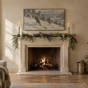 May include: A stone fireplace mantel features a green pine garland and two tall candlesticks. Above the mantel hangs a framed landscape painting of two skiers on a snowy mountain. A fire burns in the hearth, and a Christmas tree stands nearby.