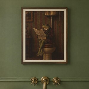 May include: A framed wall art print featuring an anthropomorphic frog wearing a dark jacket, sitting on a toilet while reading a newspaper. The scene is set in a dimly lit room with striped wallpaper, presented in a dark wood frame.