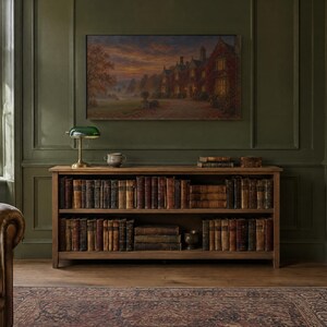 Op de afbeelding: Een lage houten boekenplank met twee niveaus gevuld met antieke boeken met lederen kaft. Bovenop staan een groene glazen bankierslamp, een kleine keramische kom en een stapel boeken. Een groot landschapsschilderij hangt aan de olijfgroene muur.