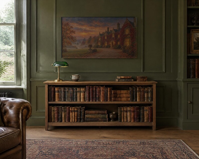 Op de afbeelding: Een lage houten boekenplank met twee niveaus gevuld met antieke boeken met lederen kaft. Bovenop staan een groene glazen bankierslamp, een kleine keramische kom en een stapel boeken. Een groot landschapsschilderij hangt aan de olijfgroene muur.