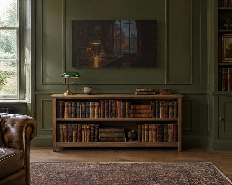 K&ouml;nnte beinhalten: Ein h&ouml;lzernes B&uuml;cherregal mit zwei Ebenen voller antiker B&uuml;cher, gekr&ouml;nt von einer gr&uuml;nen Bankerlampe aus Glas und einer kleinen Keramikschale. &Uuml;ber dem Regal zeigt ein Fernseher ein Gem&auml;lde eines Bibliotheksinnenraums.
