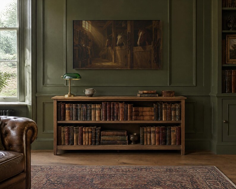 May include: A wooden bookcase with two shelves filled with antique books, topped with a green glass banker's lamp and a small ceramic bowl. Above the bookcase hangs a large rectangular painting depicting horses in a stable.