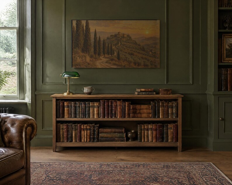 Puede incluir: Una estanter&iacute;a de madera con dos niveles llenos de libros antiguos, situada contra una pared con paneles de color verde oscuro. Sobre la estanter&iacute;a cuelga un cuadro de un paisaje con cipreses y una l&aacute;mpara de banquero verde.