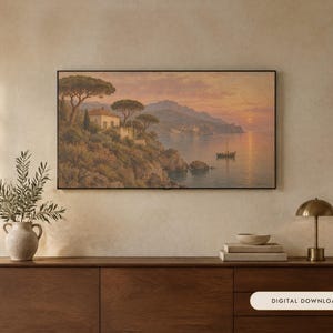 Op de afbeelding: Een rechthoekig ingelijst landschapsschilderij toont een kustsc&egrave;ne bij zonsondergang met een huis op een klif, bomen en een kleine boot op het water. Het kunstwerk hangt aan een muur boven een houten dressoir met een plant en lamp.