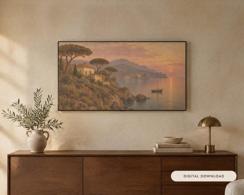 Op de afbeelding: Een rechthoekig ingelijst landschapsschilderij toont een kustsc&egrave;ne bij zonsondergang met een huis op een klif, bomen en een kleine boot op het water. Het kunstwerk hangt aan een muur boven een houten dressoir met een plant en lamp.